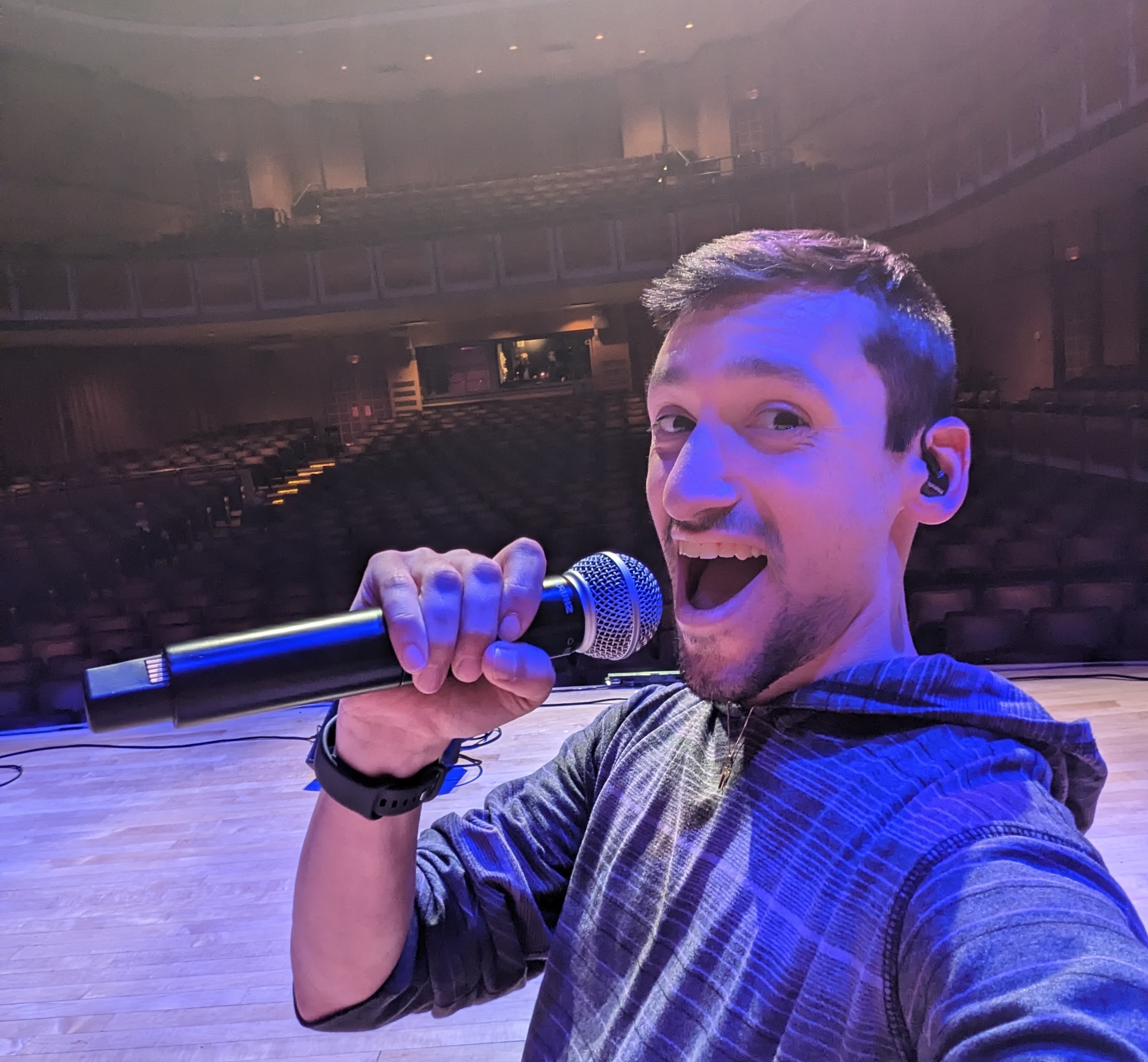 Cory Hecht holding a microphone on stage in an empty theatre