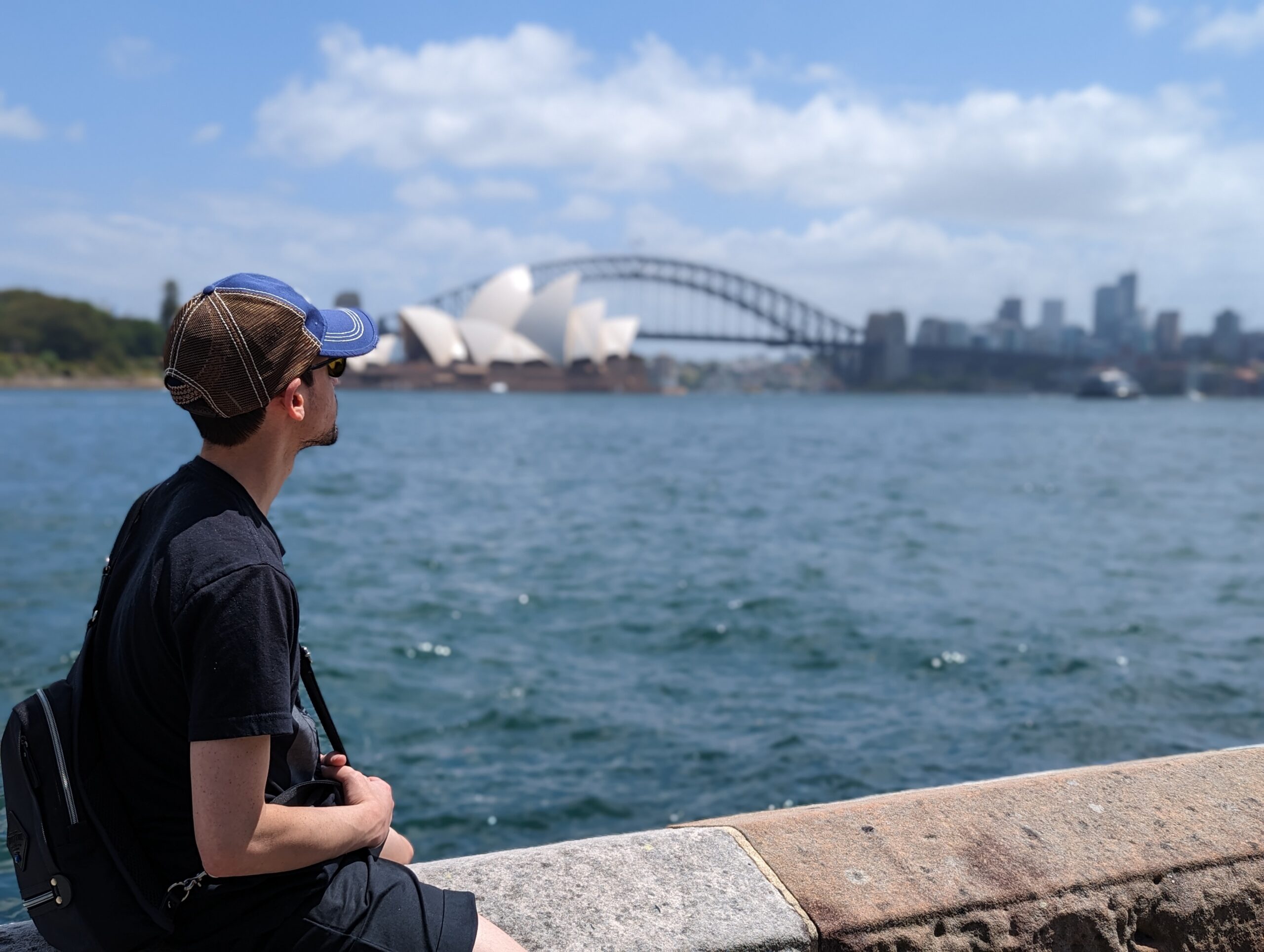 Cory Hecht sitting by Sydney Harbour with the Opera House and Harbour Bridge behind him