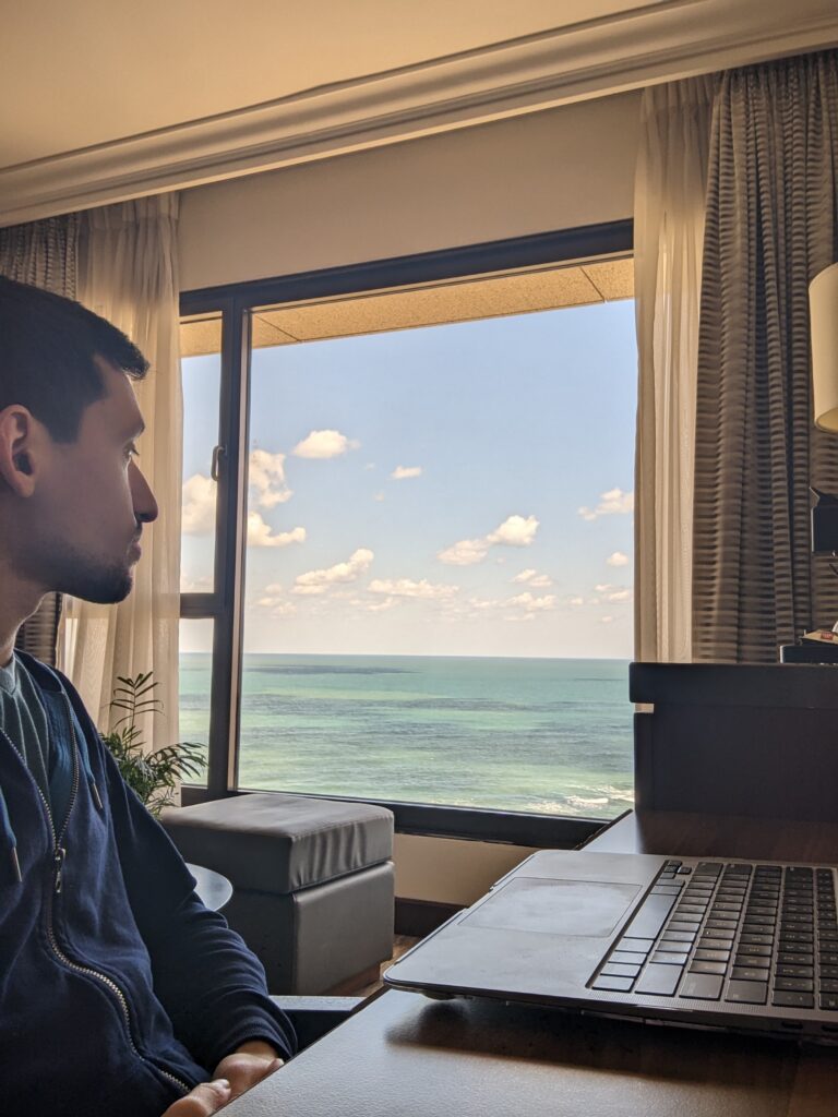 Cory Hecht working on his laptop by a window overlooking the ocean